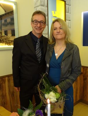 Ein Mann und eine Frau stehen eng beieinander, lächeln und halten einen Blumenstrauß. Der Mann trägt einen schwarzen Anzug und eine Brille. Die Frau trägt ein blaues Kleid und eine Halskette. Hinter ihnen ist eine Wand mit einem Bilderrahmen.