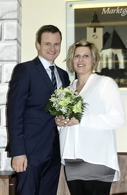 Ein Mann und eine Frau stehen lächelnd eng beieinander, die Frau hält einen Blumenstrauß. Sie sind festlich gekleidet, und eine Kirche ist im Hintergrund zu sehen.