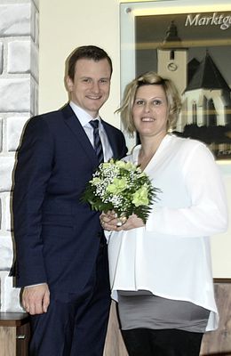 Ein Mann und eine Frau stehen lächelnd eng beieinander, die Frau hält einen Blumenstrauß. Sie sind festlich gekleidet, und eine Kirche ist im Hintergrund zu sehen.