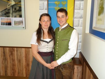 Ein lächelndes Paar in traditioneller deutscher Kleidung, das eng beieinander steht vor einer Wand mit einer Flagge.