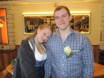 Ein junges Paar steht eng beieinander und lächelt, mit einer Kirche im Hintergrund. Der Mann trägt ein blau-weiß kariertes Hemd mit einer weißen Rose in der Tasche.