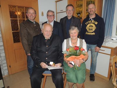 Eine Gruppe von fünf Personen steht und sitzt in einem Raum und posiert für ein Foto. Ein älterer Mann sitzt auf einem Stuhl und hält ein Geschenk und ein Papier. Eine Frau steht neben ihm und hält einen Blumenstrauß. Hinter ihnen stehen drei Männer.