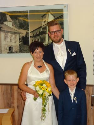 Ein frisch verheiratetes Paar und ein junger Junge stehen vor einer Wand mit einem Bild eines Gebäudes. Die Frau hält einen Blumenstrauß.