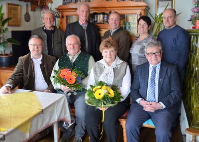 Eine Gruppe von Menschen, darunter eine Frau mit Blumen, posiert für ein Foto in einem Raum mit Holzmöbeln und einem Tisch.