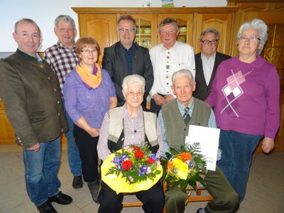 Eine Gruppe älterer Menschen, darunter zwei sitzende Personen mit Blumensträußen, versammelt sich in einem Raum mit Holzschränken und einem gefliesten Boden.