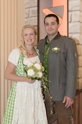Ein lächelndes Paar in traditioneller deutscher Kleidung, das eng beieinander steht, wobei die Frau einen Strauß weißer Blumen hält.
