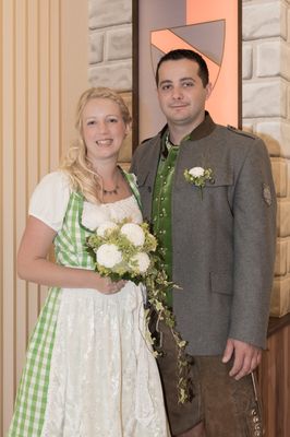 Ein lächelndes Paar in traditioneller deutscher Kleidung, das eng beieinander steht, wobei die Frau einen Strauß weißer Blumen hält.