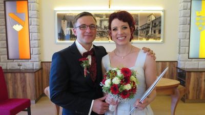 Ein frisch verheiratetes Paar steht eng beieinander und lächelt in die Kamera. Der Mann trägt einen schwarzen Anzug mit einer roten Fliege und einer Ansteckblume. Die Frau hält einen Strauß roter und weißer Blumen und trägt ein weißes Kleid. Sie stehen vor einer Wand mit einem Bilderrahmen.
