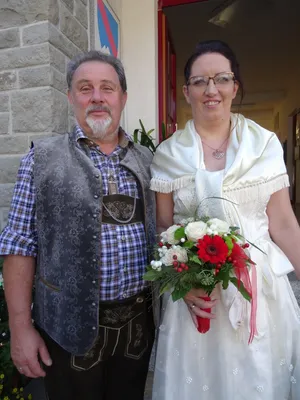 Ein Mann und eine Frau stehen lächelnd nebeneinander, der Mann in traditioneller deutscher Kleidung und die Frau in einem weißen Hochzeitskleid, die einen Blumenstrauß hält.