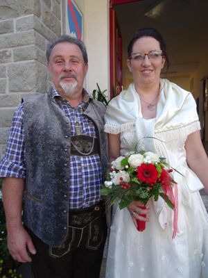 Ein Mann und eine Frau stehen lächelnd nebeneinander, der Mann in traditioneller deutscher Kleidung und die Frau in einem weißen Hochzeitskleid, die einen Blumenstrauß hält.