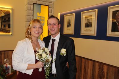 Ein Mann und eine Frau in formeller Kleidung lächeln und posieren für ein Foto vor einer Wand mit gerahmten Porträts.