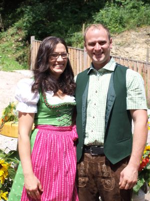 Ein Mann und eine Frau in traditioneller deutscher Kleidung stehen eng beieinander, lächeln, mit einem Holzzaun und Grünpflanzen im Hintergrund.