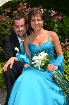 Ein Mann und eine Frau in formeller Kleidung sitzen zusammen, lächeln, die Frau hält einen weißen Blumenstrauß. Sie sind von grünem Laub umgeben.