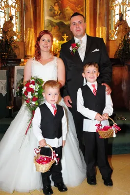 Ein frisch verheiratetes Paar steht mit ihren beiden kleinen Söhnen, alle formell gekleidet, in einer Kirche. Die Braut hält einen Blumenstrauß und die Jungen tragen kleine Körbe.