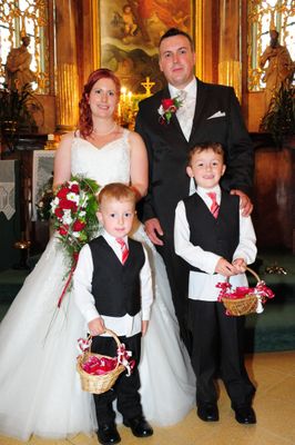 Ein frisch verheiratetes Paar steht mit ihren beiden kleinen Söhnen, alle formell gekleidet, in einer Kirche. Die Braut hält einen Blumenstrauß und die Jungen tragen kleine Körbe.