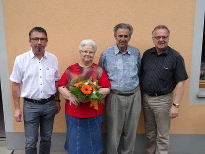Vier Personen stehen vor einer Wand und lächeln für die Kamera. Die Frau in der Mitte hält einen Blumenstrauß.