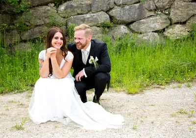 Ein frisch verheiratetes Paar, die Braut in einem weißen Kleid und der Bräutigam in einem schwarzen Anzug, sitzt auf einem Kiesweg, lächelt und posiert für ein Foto.
