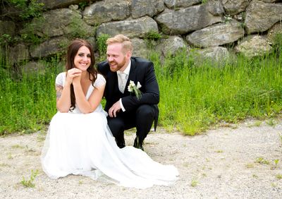 Ein frisch verheiratetes Paar, die Braut in einem weißen Kleid und der Bräutigam in einem schwarzen Anzug, sitzt auf einem Kiesweg, lächelt und posiert für ein Foto.