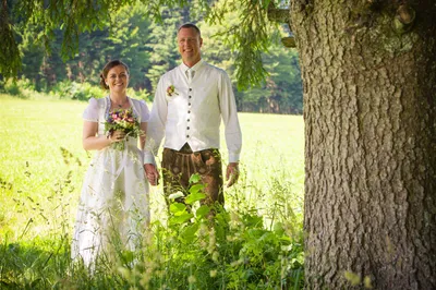 Ein frisch verheiratetes Paar steht in einem Feld, die Braut hält einen Blumenstrauß und der Bräutigam lächelt, beide in traditioneller Kleidung.