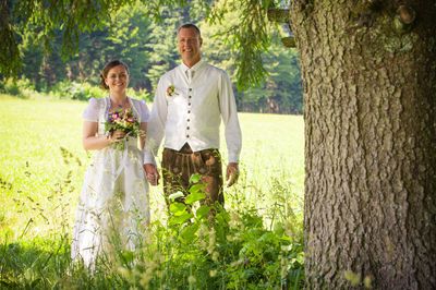 Ein frisch verheiratetes Paar steht in einem Feld, die Braut hält einen Blumenstrauß und der Bräutigam lächelt, beide in traditioneller Kleidung.