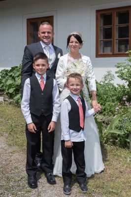 Eine Familie aus vier Personen, bestehend aus einem Mann, einer Frau und zwei Jungen, steht vor einem Haus mit Garten. Die Frau trägt einen weißen Mantel und hält einen Blumenstrauß. Die Jungen sind in schwarze Weste und rote Krawatten gekleidet.