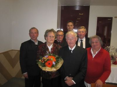 Eine Gruppe älterer Personen, darunter eine Frau mit einem Blumenstrauß, steht in einem Raum zusammen.