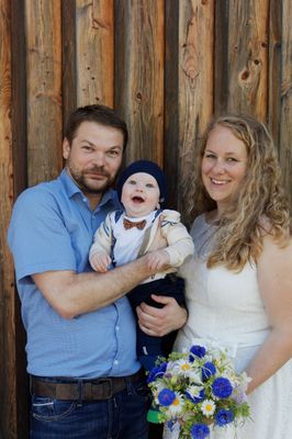 Eine Familie aus drei Personen, ein Mann, eine Frau und ein Baby, lächelt und posiert für ein Foto vor einer Holzwand.