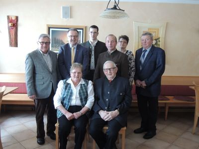 Eine Gruppe von Menschen, darunter Männer und Frauen, stehen und sitzen in einem Raum, wahrscheinlich für ein Gruppenfoto. Sie tragen alle formelle Kleidung.