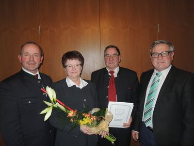 Vier Menschen stehen zusammen und lächeln für ein Foto. Eine Frau hält einen Blumenstrauß und ein Zertifikat. Die Männer tragen Anzüge und Krawatten.
