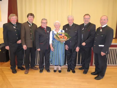 Eine Gruppe formell gekleideter Männer und eine ältere Frau mit einem Blumenstrauß posieren für ein Foto auf einem Holzboden.
