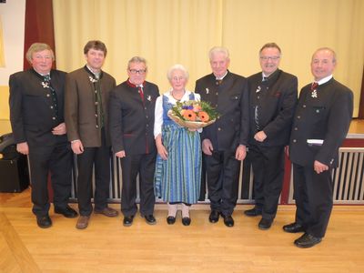 Eine Gruppe formell gekleideter Männer und eine ältere Frau mit einem Blumenstrauß posieren für ein Foto auf einem Holzboden.