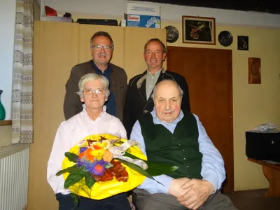 Vier Männer in einem Raum mit einer Frau, die einen Blumenstrauß hält. Zwei Männer stehen hinter ihnen.