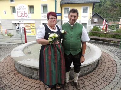 Ein Mann und eine Frau in traditioneller deutscher Kleidung stehen vor einem Brunnen und lächeln für ein Foto. Hinter ihnen steht ein Gebäude mit einem Schild, das Cafe Backerei besagt.