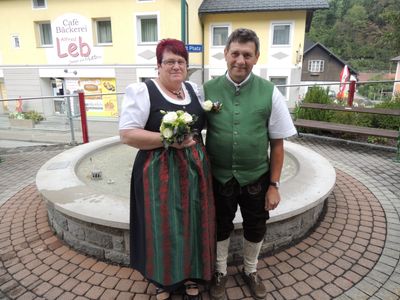 Ein Mann und eine Frau in traditioneller deutscher Kleidung stehen vor einem Brunnen und lächeln für ein Foto. Hinter ihnen steht ein Gebäude mit einem Schild, das Cafe Backerei besagt.