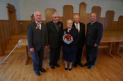 Fünf Personen, vier Männer und eine ältere Frau, stehen in einem Raum zusammen. Die Frau hält einen Strauß roter Rosen. Sie sind alle in formeller Kleidung gekleidet. Hinter ihnen befindet sich eine Holzstruktur.