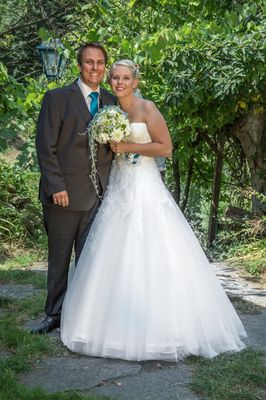 Ein frisch verheiratetes Paar steht in einem Garten, die Braut in einem weißen Kleid und der Bräutigam in einem dunklen Anzug.