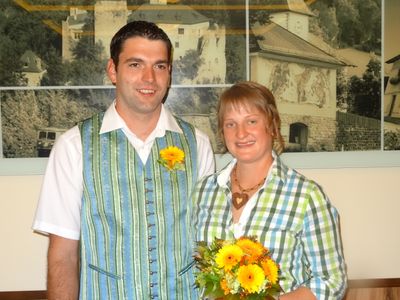 Ein Mann und eine Frau stehen eng beieinander und lächeln für ein Foto. Der Mann trägt eine blaue Weste mit einer gelben Blume, und die Frau hält einen Strauß gelber Blumen. Im Hintergrund ist eine landschaftliche Kulisse.