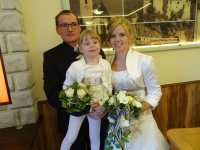 Ein Mann und eine Frau in formeller Kleidung stehen mit einem jungen Mädchen, das Blumen hält, und lächeln für ein Foto.