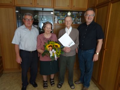 Vier Personen stehen in einem Raum, zwei Männer und zwei Frauen. Der ältere Mann hält einen Blumenstrauß und ein Buch. Die Frau neben ihm hält den Strauß. Die anderen beiden Männer stehen auf beiden Seiten.