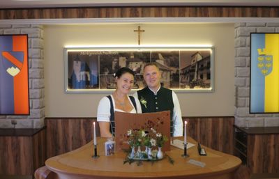 Ein lächelndes Paar steht an einem Schreibtisch mit einer Blumenarrangement, Kerzen und einem gerahmten Foto über ihnen. Der Mann trägt eine Weste, und die Frau trägt eine Halskette.