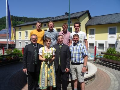 Eine Gruppe von Menschen, darunter ein Paar in traditioneller Kleidung, steht vor einem gelben Gebäude mit einem Brunnen im Vordergrund.