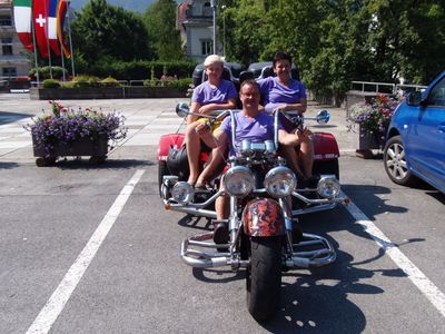 Drei Personen in passenden lila T-Shirts fahren auf einem roten Trike-Motorrad auf einem Parkplatz.