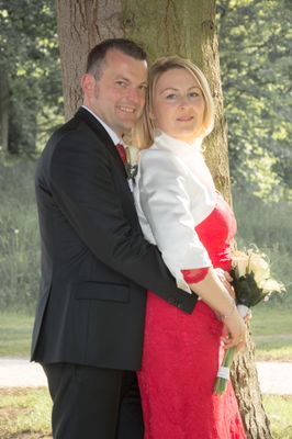 Ein Mann und eine Frau stehen eng beieinander und lächeln in einem Park mit einem Baum im Hintergrund. Die Frau hält einen Blumenstrauß.