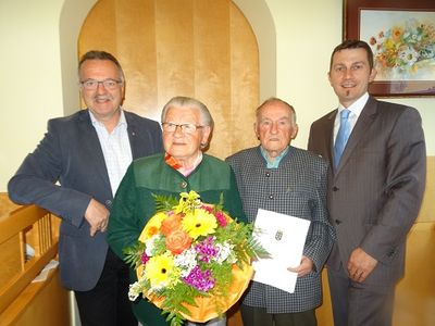 Drei Männer in Anzügen stehen hinter einer älteren Frau, die einen Blumenstrauß und ein Zertifikat in der Hand hält.
