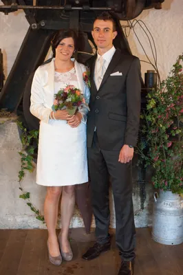 Eine lächelnde Braut in einem weißen Kleid und ein Bräutigam in einem schwarzen Anzug stehen zusammen und halten Händchen. Sie sind von Grünpflanzen und einer dekorativen Pflanze umgeben.