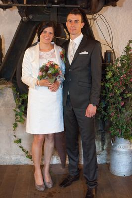 Eine lächelnde Braut in einem weißen Kleid und ein Bräutigam in einem schwarzen Anzug stehen zusammen und halten Händchen. Sie sind von Grünpflanzen und einer dekorativen Pflanze umgeben.