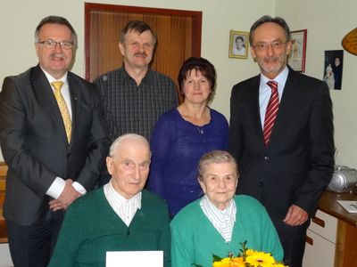 Eine Gruppe von sechs Personen, darunter zwei ältere Menschen, steht in einem Raum und lächelt für ein Foto. Das ältere Paar sitzt, während die anderen hinter ihnen stehen. Ein Blumenstrauß steht vor dem sitzenden Paar.