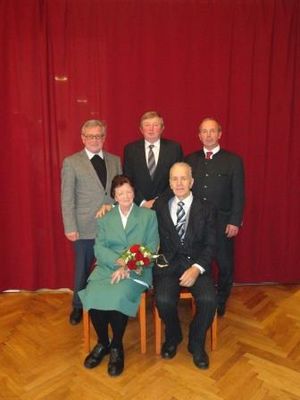 Fünf Personen in formeller Kleidung posieren für ein Foto vor einem roten Vorhang. Eine Frau in der Mitte hält einen Blumenstrauß.