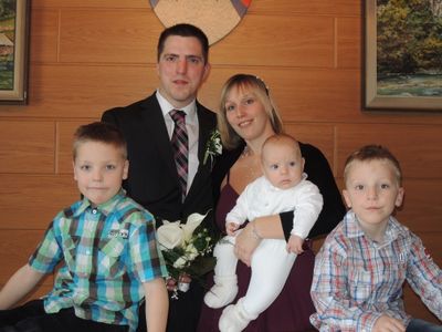 Eine Familie mit fünf Personen, darunter ein Mann im Anzug, eine Frau mit einem Baby und drei Kinder, posiert für ein Foto vor einer Holzwand.
