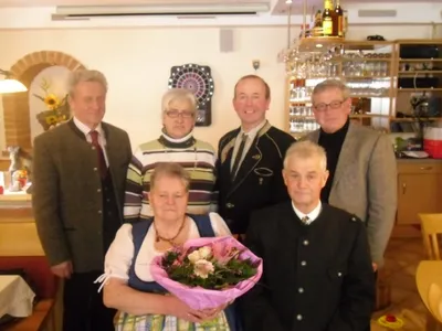 Eine Gruppe von sechs Personen steht in einem Restaurant, wobei eine Frau in der Mitte einen Blumenstrauß hält. Hinter ihnen befindet sich ein Dartboard und Regale mit Gläsern.